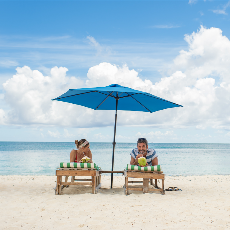 A Couple Of People Sitting On A Bench Under An Umbrella At The Beach